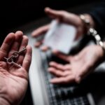 Mâini încătușate deasupra unei tastaturi de laptop, într-o cameră întunecată