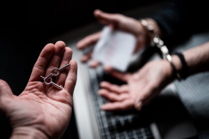 Mâini încătușate deasupra unei tastaturi de laptop, într-o cameră întunecată